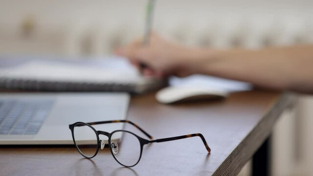 The glasses are on the work table. Against a blurred background, the guy works at a laptop and makes notes in a notebook.