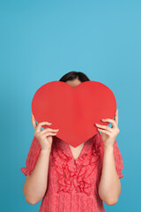 a dark-haired woman in a red dress hides her face behind a large red paper heart, isolated on a blue background.