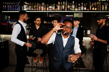 Obraz premium Stylish african american man against group of handsome retro well-dressed guys gangsters spend time at club, drinking on bar counter. Multiethnic male bachelor mafia party in restaurant.