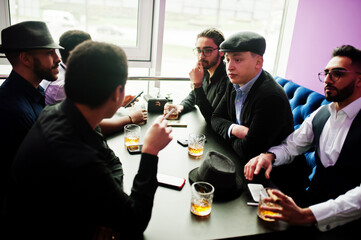 Group of handsome retro well-dressed man gangsters spend time at club, sitting and drinking whiskey. Multiethnic male bachelor mafia party in restaurant.