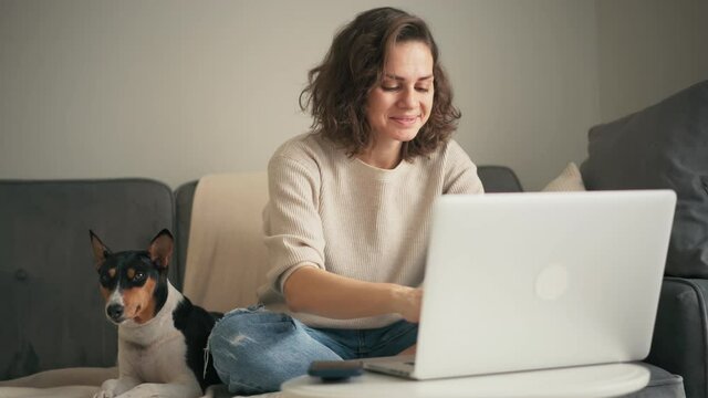 A young woman working on a laptop while sitting on a sofa in the living room with her cute basenji dog. 