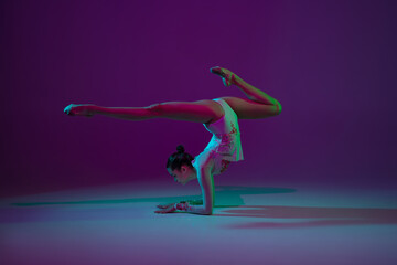 Balance. Young female athlete, rhythmic gymnastics artist dancing, training isolated on purple studio background in neon light. Beautiful girl practicing with equipment. Grace in performance.