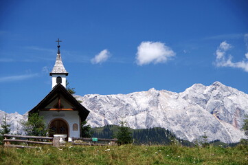 Fototapeta na wymiar Kleine Bergkapelle auf einer Bergwiese mit gewaltigem Gebirgsmassiv im Hintergrund unter blauem Himmel