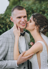 Obraz premium A beautiful bride in a white dress with a long veil kisses the cheek of a smiling groom in a gray suit in the park against a background of green tui. Wedding portrait of newlyweds in love.