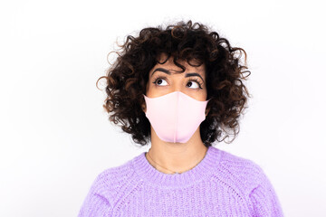 young beautiful caucasian woman wearing medical mask standing against white wall looking aside into empty space thoughtful