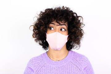 Amazed young beautiful caucasian woman wearing medical mask standing against white wall bitting lip and looking tricky to empty space.