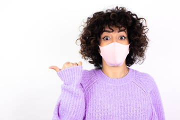 Lovely young beautiful caucasian woman wearing medical mask standing against white wall pointing aside with forefinger, showing at copy space having news about bargains