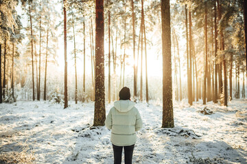 A female in winter hat and puffer standing and observing the beautiful sunset over snow-covered forest