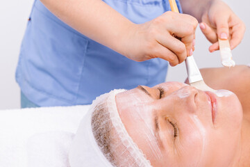 Cropped photo of a beautician applying a moisturizing face mask with a cosmetic brush to a senior woman a headband bandage on her head . Spa and beauty concept