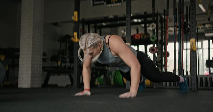 Tracking shot of blond female athlete doing squat thrust exercise during intense workout in gym