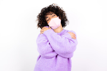 young beautiful caucasian woman wearing medical mask standing against white wall Hugging oneself...