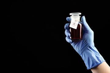 Woman in gloves holding glass bottle of poison with warning sign on black background, closeup. Space for text