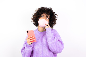 young beautiful caucasian woman wearing medical mask standing against white wall thinks deeply...
