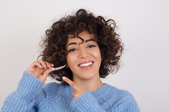 young beautiful caucasian woman wearing blue knitted sweater standing against white studio background holding an invisible aligner braces and smiling. Dental healthcare concept. 