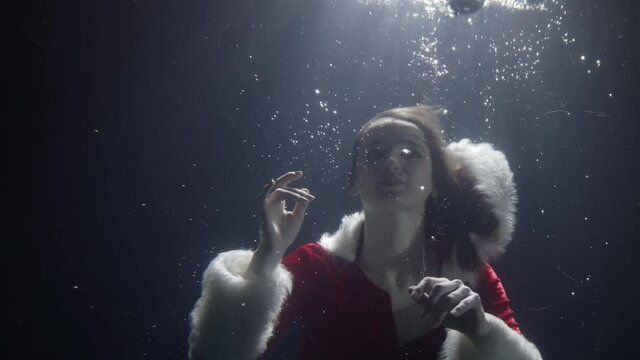 an attractive woman in red Christmas clothes with white fur is under water, holding Christmas balls in her hands, letting them float up, creating a lot of bubbles with her hands.