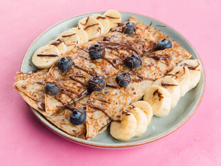 Delicious crepes filled with chocolate hazelnut spread and topped with banana and blueberries. Breakfast, brunch dessert on bright pink background, closeup