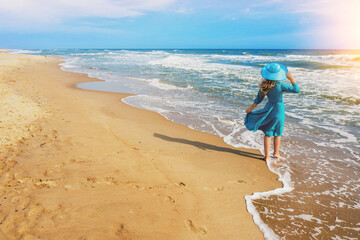 Seascape on a sunny summer day. The girl on the beach looks at the sea. Young happy girl carefree walk along the sea in blue fluttering dress and sun hat