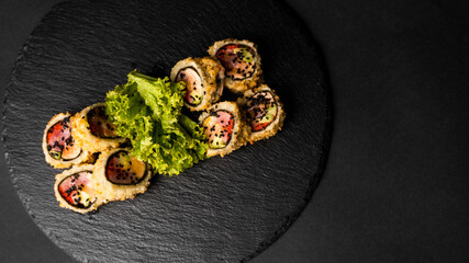 Custom sushi roll in tempura with nori, fresh salmon, tuna, avocado, masago caviar, drizzled with pineapple sauce with salad pouring as decoration on a black plate on a black table and background.
