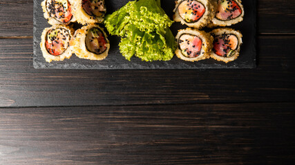 Custom sushi roll in tempura with nori, fresh salmon, tuna, avocado, masago caviar, drizzled with pineapple sauce with salad pouring as decoration on a black plate on a wooden table and background.