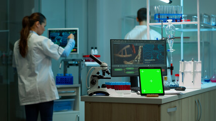 Microbiologist working on monitor in background analysing test samples with tablet with green chroma key screen placed on desk in front. Team of biotechnology scientists developing drugs.
