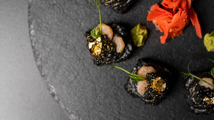 Sushi roll with nori, black rice, crab meat, cucumber, avocado, smoked salmon mousse, oar caviar, masago, shrimp cocktail and edible gold leaf with red ginger and wasabi on black table and background.