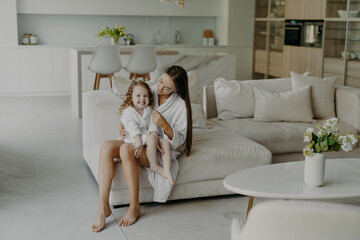Obraz na płótnie Canvas Happy adorable little hands poses on mothers legs dressed in soft white bath towel feels very glad. Mom and daughter wear domestic clothes sit on comfortable sofa in living room. Home interior