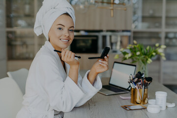 Tender lovely woman with healthy skin holds small mirror and cosmetic brush applies powder on face smiles toothily wears soft white bathrobe and towel poses against home interior. Beauty concept