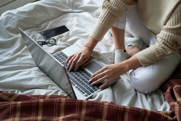 Young beautiful girl in cozy sweater in bed at home working on a laptop
