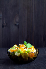 healthy salad from fruit with green mint on a wood table. Mixed fresh banana, orange, kiwi, fruit on the black background. copy space. Refreshing fruit salad. vertical