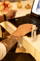 close up of a person with tattoos working on a laptop with hand sanitizer next to him. 