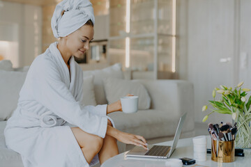 Indoor shot of female freelancer workds distantly rom home types on laptop computer dressed in bathrobe drinks coffee or tea enjoys domestic atmosphere surfs internet reads advice about skin care