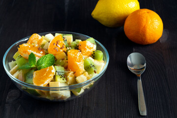 Refreshing fruit salad with spoon and lemon, orange. healthy salad from fruit with green mint on a wood table. Mixed fresh banana, orange, kiwi, fruit on the black background. copy space.