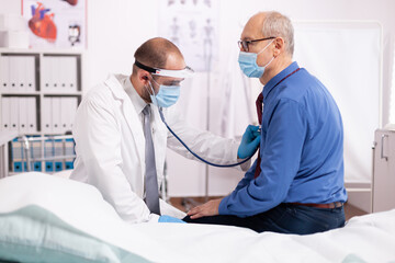 Medical practitioner wearing face mask consulting senior man in examination room during coronavirus.