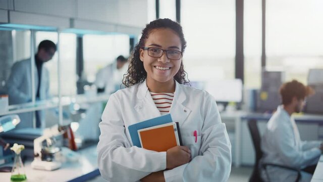 Medical Science Laboratory: Beautiful Smart Young Black Scientist Wearing White Coat and Glasses, Holds Test Books, Smiles Looking at Camera. Diverse Team of Specialists. Medium Portrait Shot