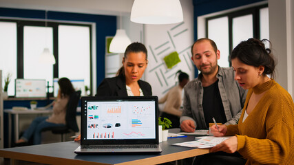 Obraz premium Team of colleagues employees cooperating sitting at desk in startup company office analysing graphs, taking notes. Business coworkers planning start up business project, brainstorming in wokrplace
