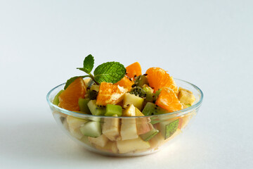 fruit salad in glass bowl on white background. Bowl of healthy salad from fruit with green mint on a white table. Mixed fresh banana, orange, kiwi, fruit. copy space. Healthy vegan lunch.