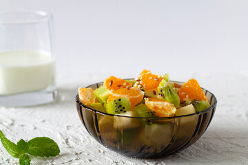 Healthy vegan lunch. Bowl of healthy salad from fruit with green mint on a white table. Mixed fresh banana, orange, kiwi, fruit. copy space. fruit salad in glass dark bowl with glass of milk on white 