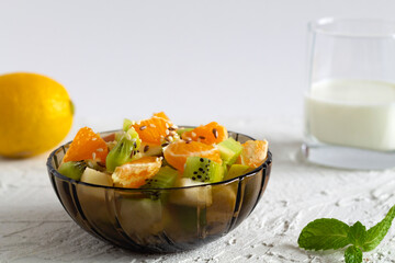 Bowl of healthy salad from fruit with green mint on a white table. Mixed fresh banana, orange, kiwi, fruit. copy space. fruit salad in glass dark bowl with glass of milk on white background.