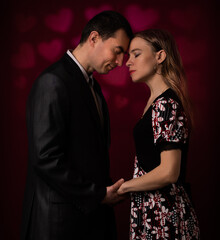 Young man and woman standing together holding hands against red wall with heart shapes