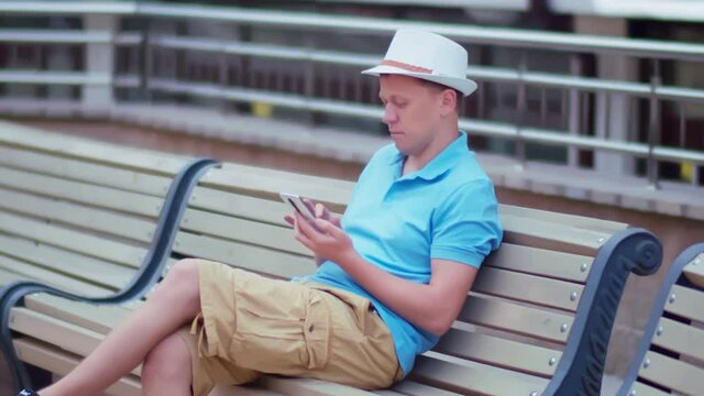 Young man in a hat sits on a city bench with a mobile phone in his hands, camera tracking