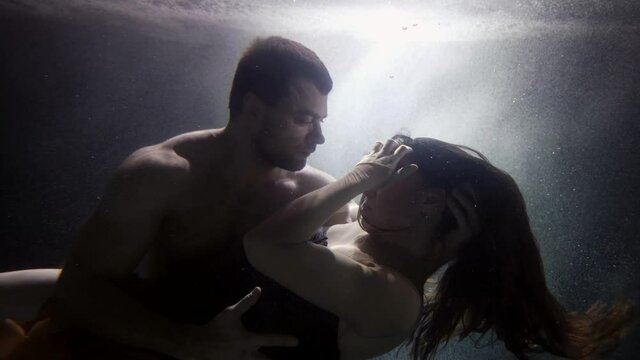 A man holds a woman under the water by the waist, passionately embraces, kisses and swims together under the water, they spin like in a fairy tale in bubbles.