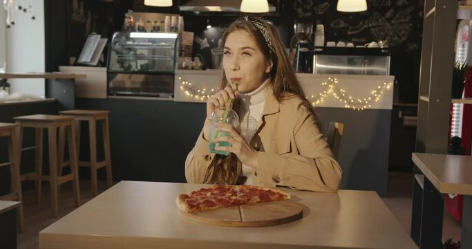 Laughing young long-haired girl sits in a cafe with leftover pizza on a wooden plank and drinks a blue cocktail through a straw