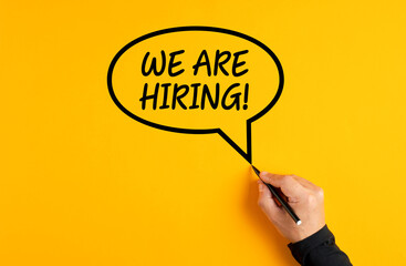 Male hand drawing a speech balloon with the message of we are hiring on yellow background. Job...