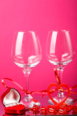 Two champagne glasses with ribbons on a pink background and a red heart-shaped box with a ring.