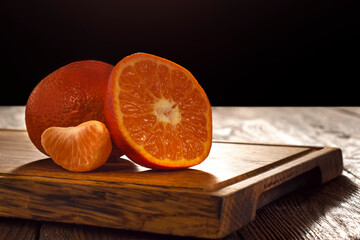 A whole and cut tangerine with a wedge lies on a wooden table in a dark vein.