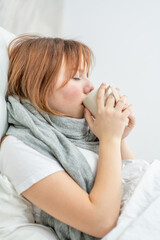 Sick girl sits on a bed at home and drinks hot tea from a mug