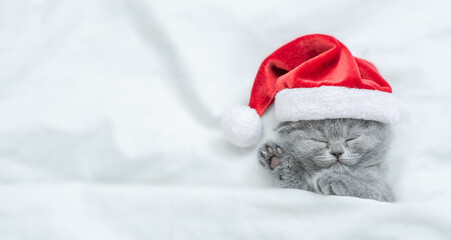 Funny kitten wearing red santa's hat sleeps under a white blanket on a bed. Top down view. Empty...