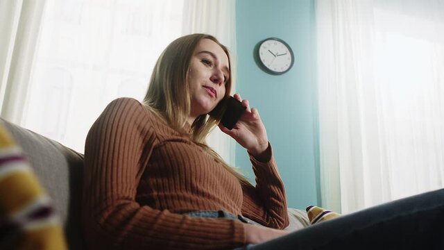 Young Lady in a brown sweater and blue jeans sits on the couch in front of the window, carefree talking on a cell phone and twisting her hair. Lower angle