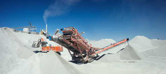 Mobile crusher machine and excavator on the background of crushed stone hills and blue sky, panorama.