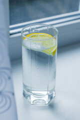 A glass of water with a slice of lemon stands on a white windowsill by the window behind a white tulle